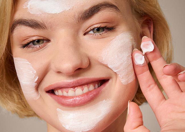 Eine lächelnde Person trägt weiße Creme Maske auf das Gesicht auf.