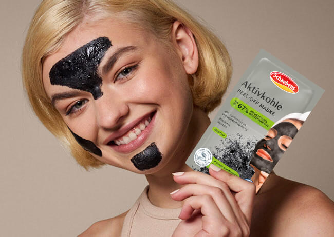 Blonde Frau mit schwarzer Gesichtsmaske lächelt und hält eine Packung Aktivkohle-Peel-off-Maske in der Hand.
