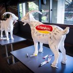IMG_0208 Zwei weiße Ziegenfiguren mit Unterschriften stehen auf Tischen, umgeben von farbigen Stiften.