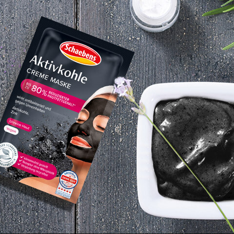 Eine Schaebens Aktivkohle-Creme-Maske liegt neben einer Schale mit schwarzer Creme auf grauem Holztisch.