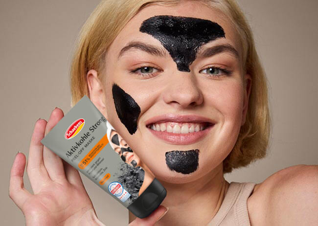 Frau mit schwarzer Gesichtsmaske lächelt, hält eine Tube mit Peeling-Maske in der Hand.