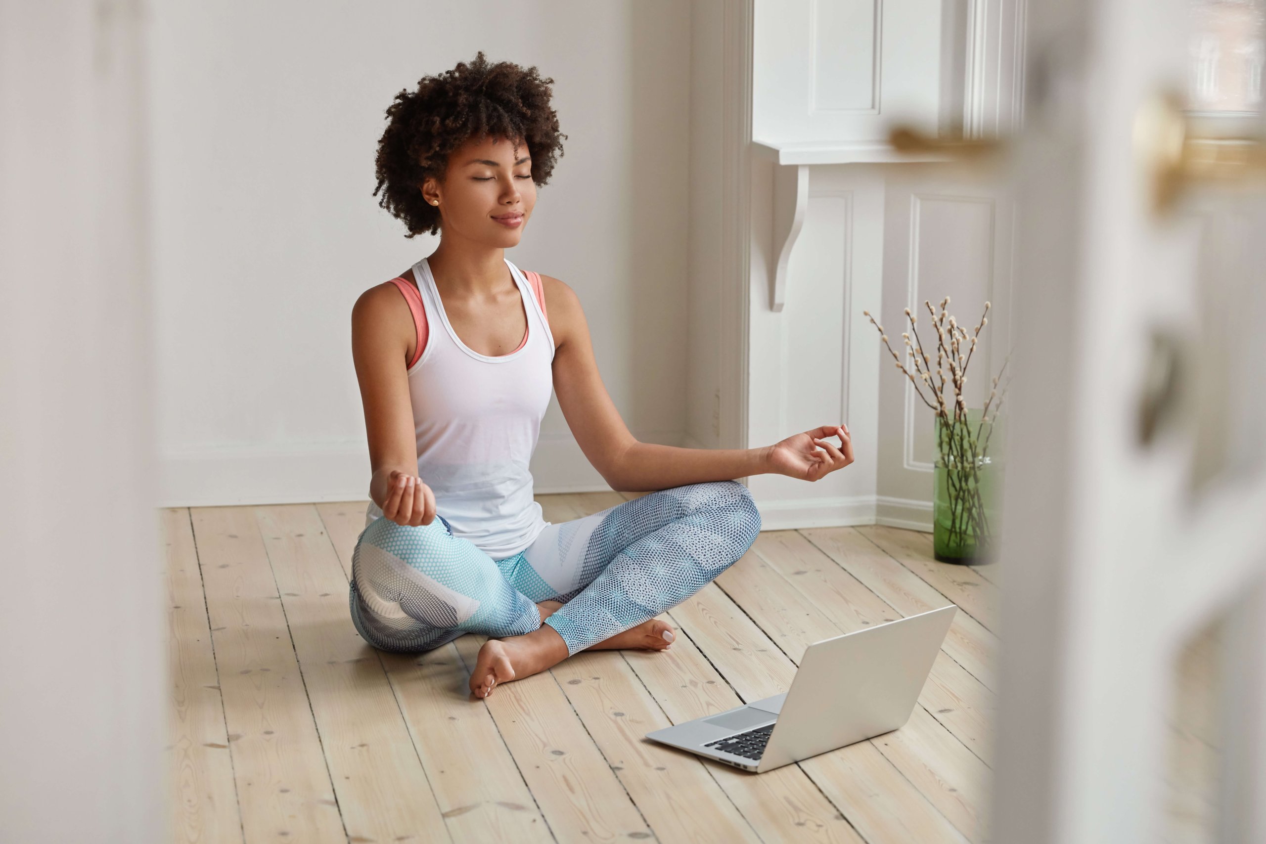 Relaxed dark skinned woman with sporty body, sits in zen pose, keeps legs crossed, watches yoga lessons on laptop computer using internet, meditates on floor in empty room. Wellness concept. yoga-laptop-work-life-balance-worklifebalance-relax-chill-tipps