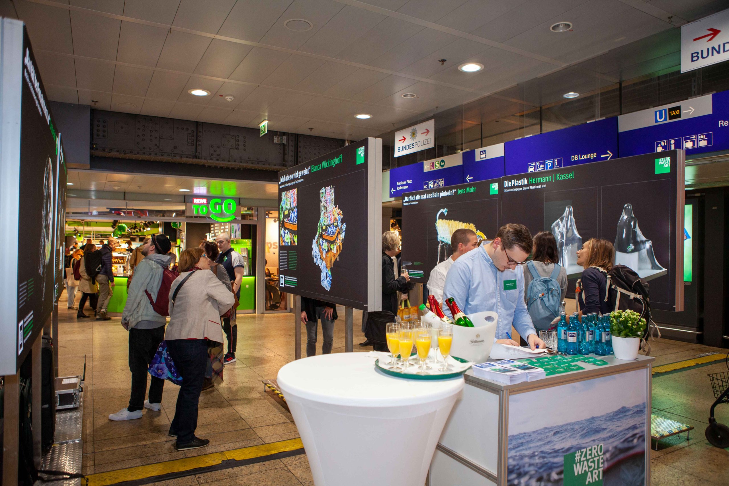 ZEROWASTEART_8 Menschen betrachten Kunsttafeln in einem Bahnhof, im Vordergrund ein Tisch mit Getränken.