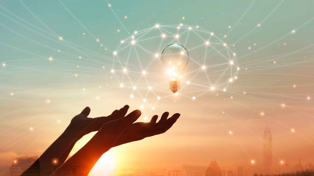 Hände heben sich zu einer leuchtenden Glühbirne in einem Netz aus Lichtpunkten vor einem Sonnenuntergang.