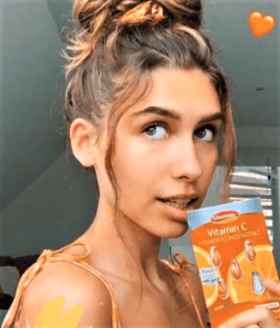 Eine Frau hält eine Schaebens Packung Vitamin C Konzentrat, trägt einen Dutt und lächelt leicht, mit einem Herz-Emoji im Hintergrund.