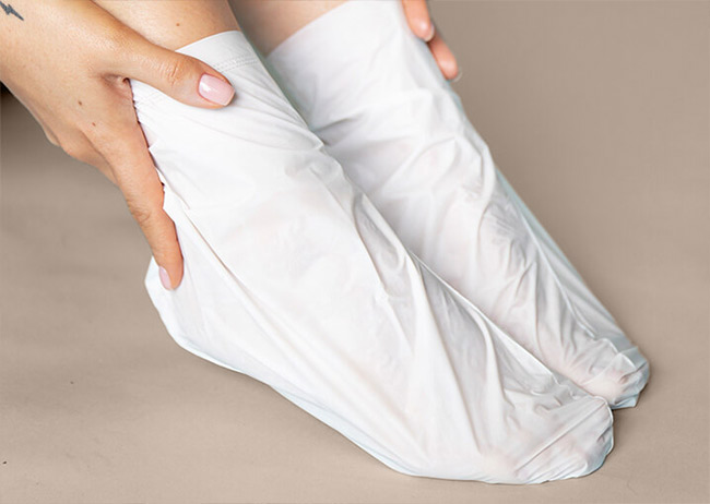 Zwei Füße in weißen Fußmasken auf beigem Hintergrund, Hände berühren die Knöchel.