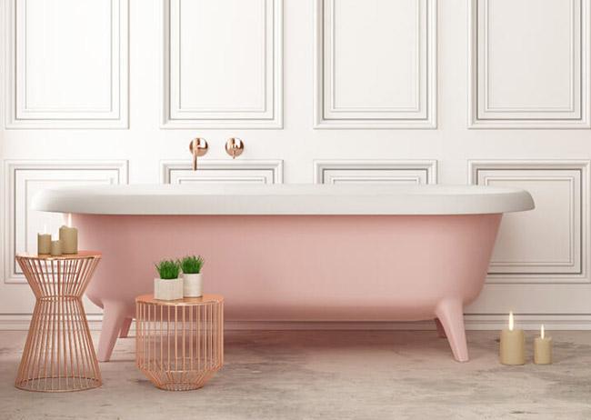 Rosa Badewanne vor weißer Wand, umgeben von Kupfertischen mit Kerzen und Pflanzen.