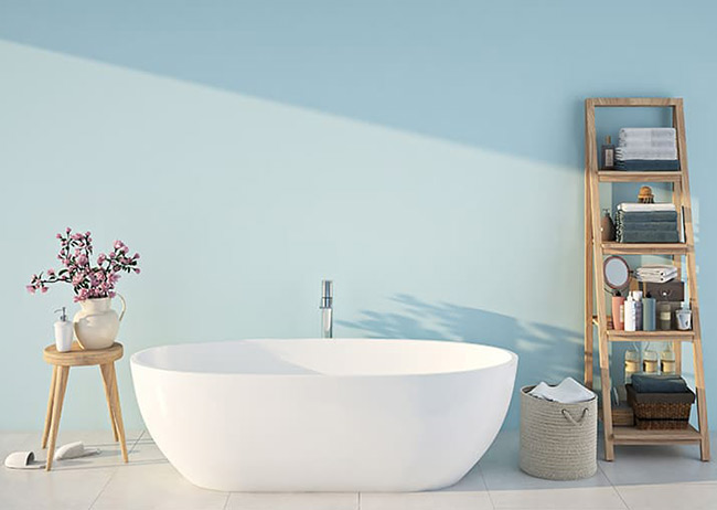 Weiße Badewanne vor blauer Wand, daneben Holzregal mit Handtüchern, Korb und Blumen auf Hocker.