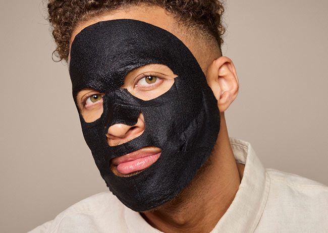 Mann mit schwarzer Gesichtsmaske, lockigem Haar, trägt ein helles Hemd, schaut in die Kamera.