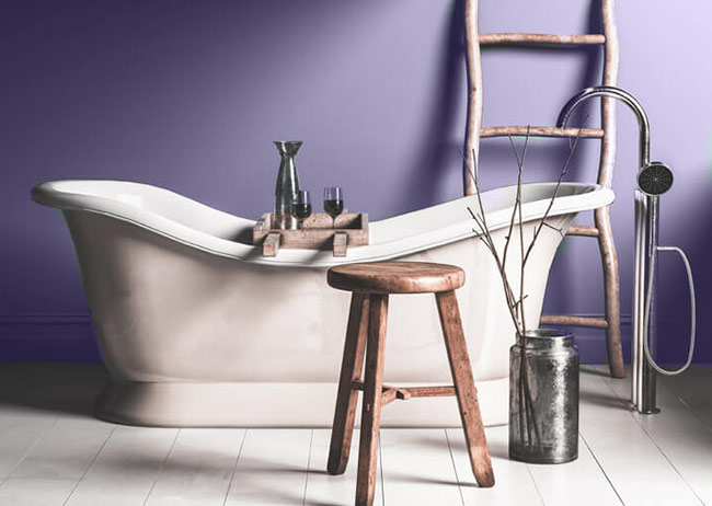 Freistehende Badewanne vor lila Wand, Holzleiter, Hocker und Vase mit Zweigen.