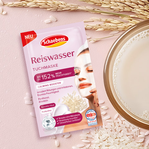 Packung einer Reiswasser Tuchmaske von Schaebens auf rosa Hintergrund, umgeben von Reiskörnern und einer Schale mit Flüssigkeit.