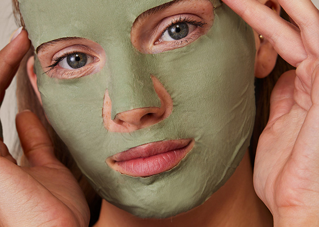 Person mit grüner Gesichtsmaske, Schaebens Tonerde Tuchmaske, Hände am Gesicht, schaut direkt in die Kamera.