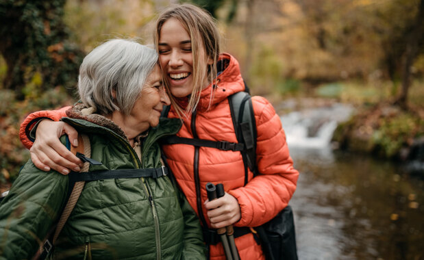 Zwei lächelnde Frauen in Winterjacken umarmen sich vor einem Bach in einer herbstlichen Landschaft.