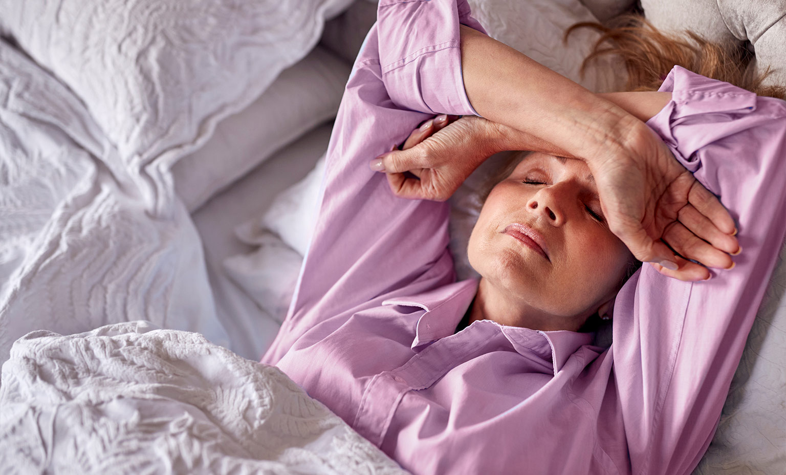 Frau in lila Hemd liegt im Bett, Arme über Stirn, Augen geschlossen, weiße Bettwäsche.