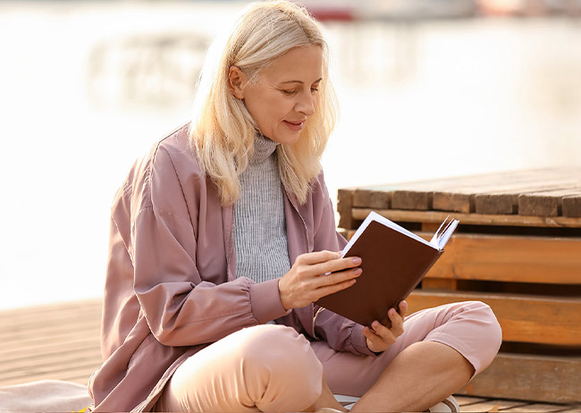 Eine Frau in rosa Kleidung liest draußen ein Buch, sitzt entspannt auf einem Holzsteg.
