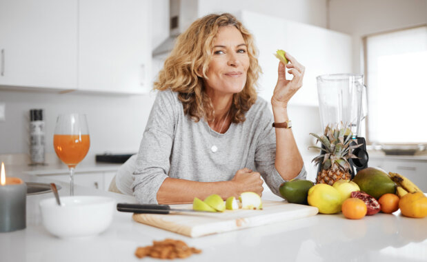 Frau in Küche mit geschnittenem Apfel, Obst, Saft und Kerze auf weißer Arbeitsfläche.