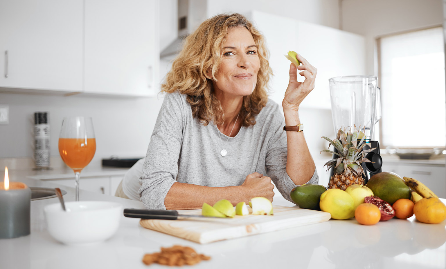 Frau in Küche mit geschnittenem Apfel, Obst, Saft und Kerze auf weißer Arbeitsfläche.