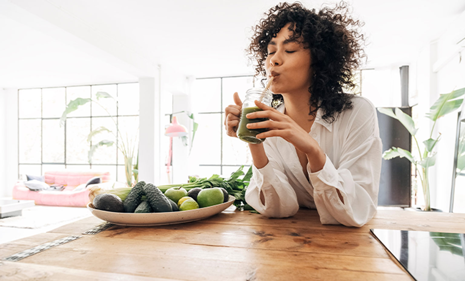 Eine Frau trinkt einen grünen Smoothie am Holztisch mit Avocados und Limetten.