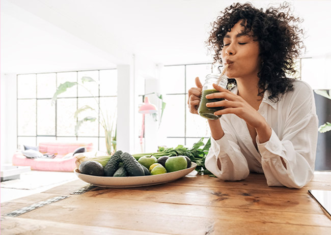 Eine Frau trinkt grünen Smoothie an einem Tisch mit Avocados und Limetten in einem hellen Raum.