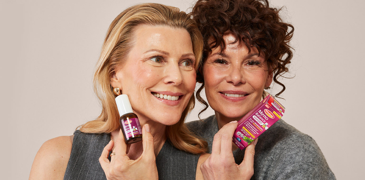Zwei lächelnde Frauen halten Schaebens Meno Balance Sofort Spray in der Hand, vor neutralem Hintergrund.